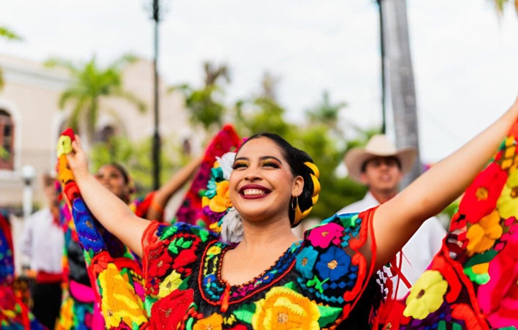 Smiling woman in vibrant, floral Mexican folk dress dancing at an outdoor cultural festival.
