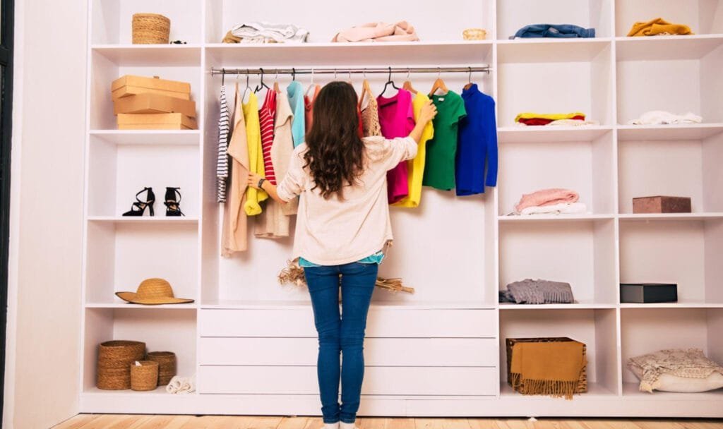 Woman organizes a white walk-in closet with a mix of colorful vintage and modern clothing on a hanging rack.