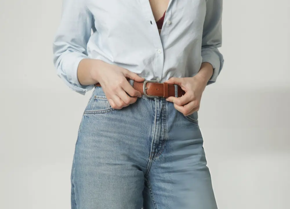 Woman fastening a brown leather belt on high-waisted jeans, showing weight loss progress.