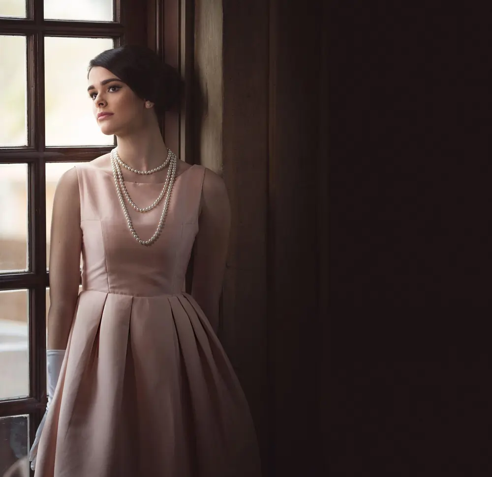 Woman in a vintage pink dress and pearls looking out a window.